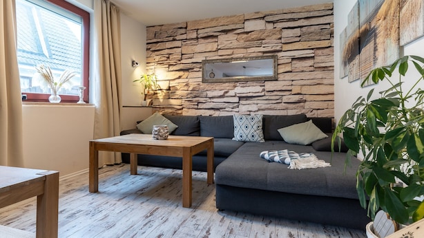 A cozy living room with a stone wall, a dark grey sofa, a wooden coffee table, and a large window with curtains. - Moderne und Komfortable Ferienwohnung in Busum (Büsum)