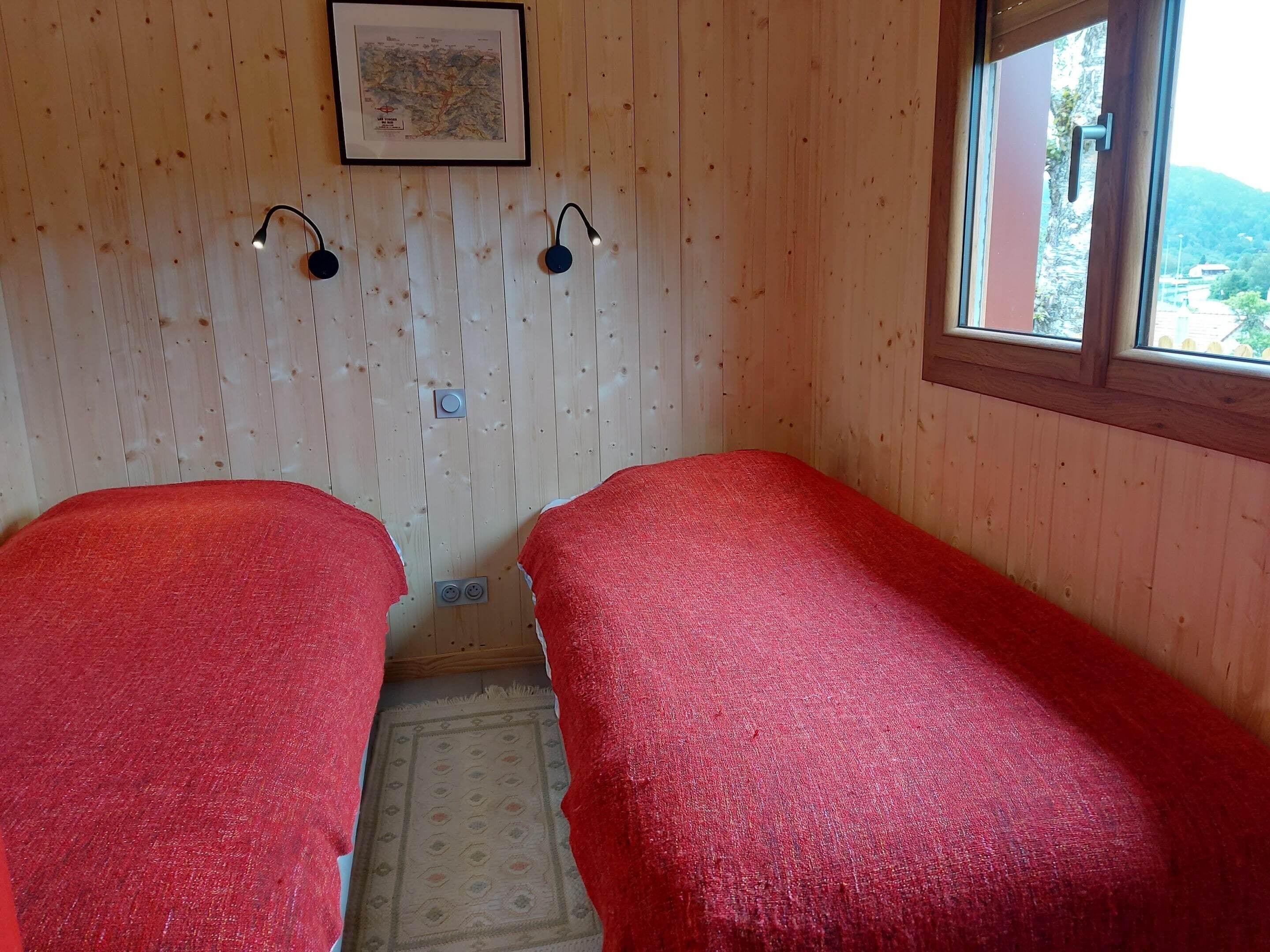 Une chambre avec deux lits simples recouverts de couvertures rouges, un mur en bois orné d’une carte encadrée, deux lampes de lecture fixées au mur et une fenêtre donnant sur des collines.