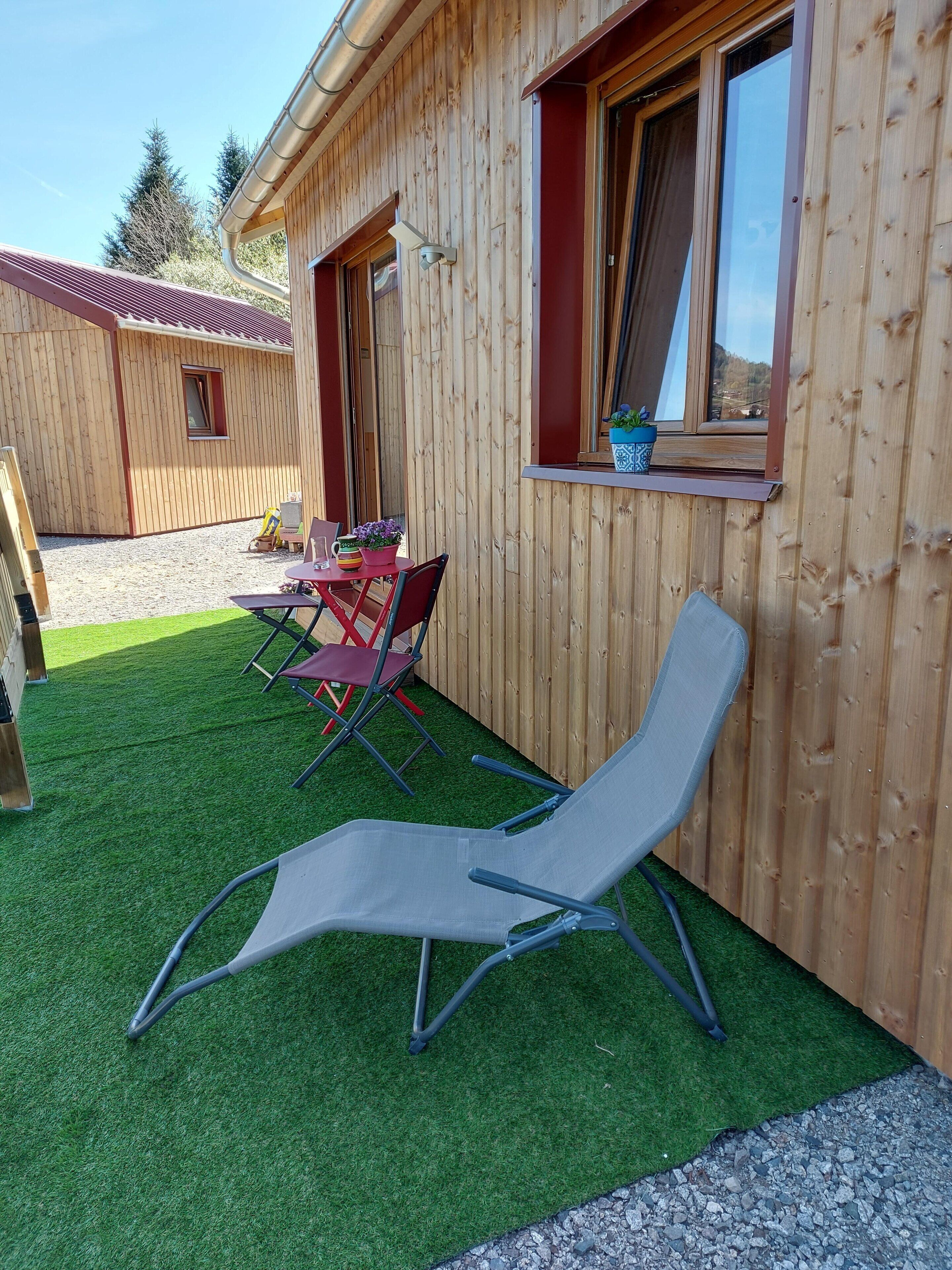 A wooden cabin with a patio area featuring artificial grass, a table with chairs, and a lounge chair.