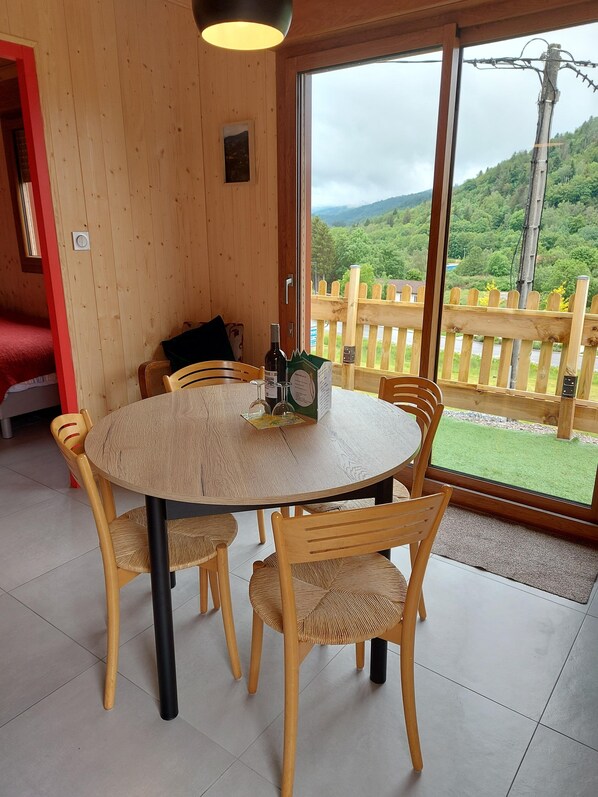 A wooden interior with a round table and chairs, a window with a view of a green landscape, and a red door. - 2 Chalets for 4 People (Saint-Maurice-sur-Moselle)