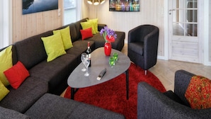 Un salon moderne avec un canapé gris foncé, des coussins rouges et jaunes, une table basse ronde sur laquelle se trouvent une bouteille de vin, des verres et une télécommande. On peut également apercevoir un fauteuil noir et un tapis rouge.