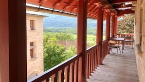 Un espace extérieur abrité, doté d’une terrasse en bois, d’une table et de chaises, qui offre une vue sur un bâtiment et un paysage verdoyant, avec des montagnes à l’horizon.