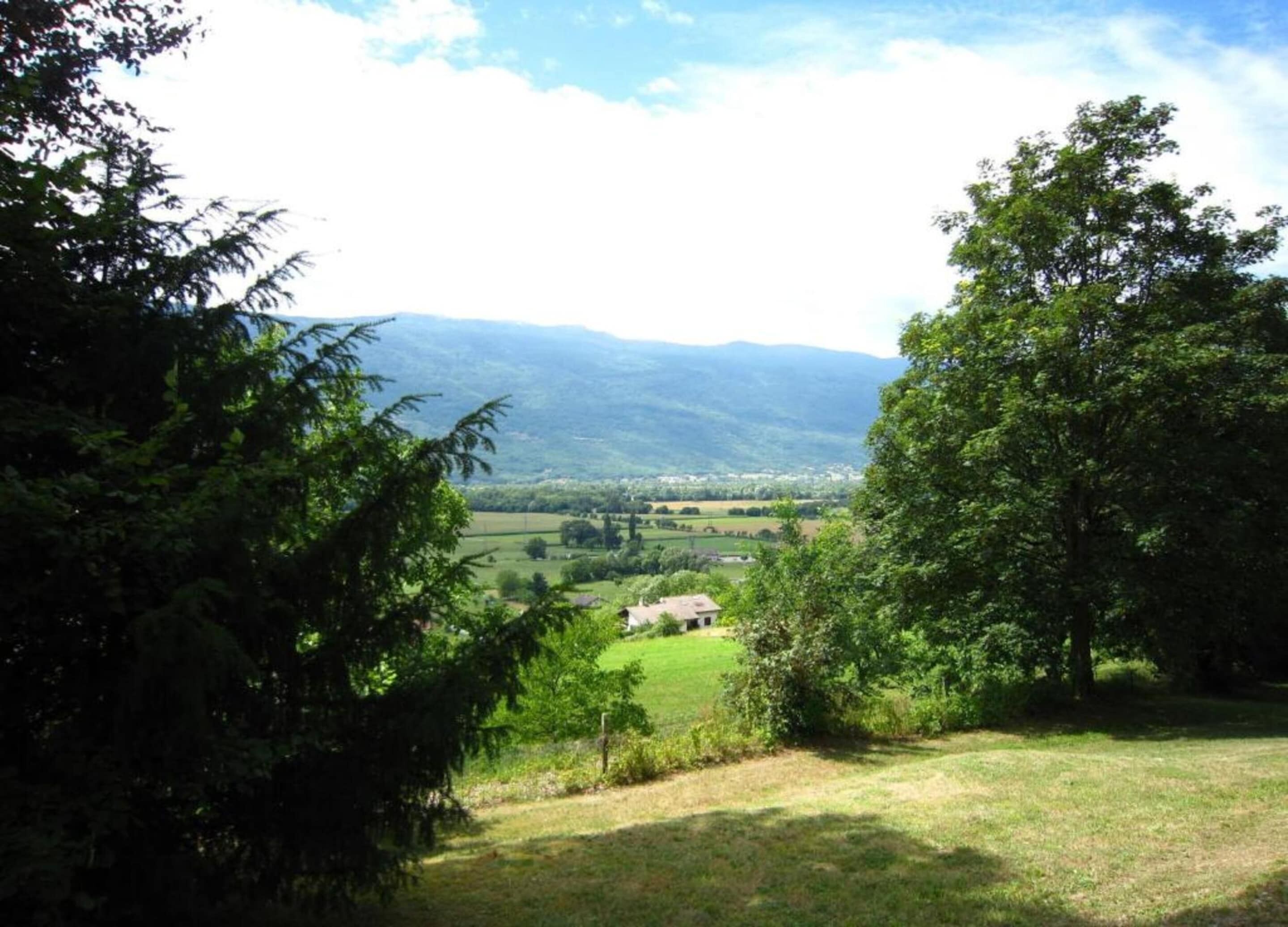 Ländliche Landschaft mit einem Haus, umgeben von Bäumen und Feldern, mit Blick auf ferne Hügel.