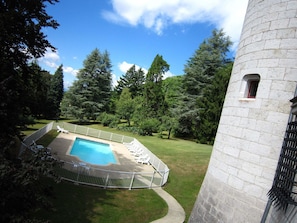 A castle-like structure with a swimming pool surrounded by a fence, trees, and a clear sky.