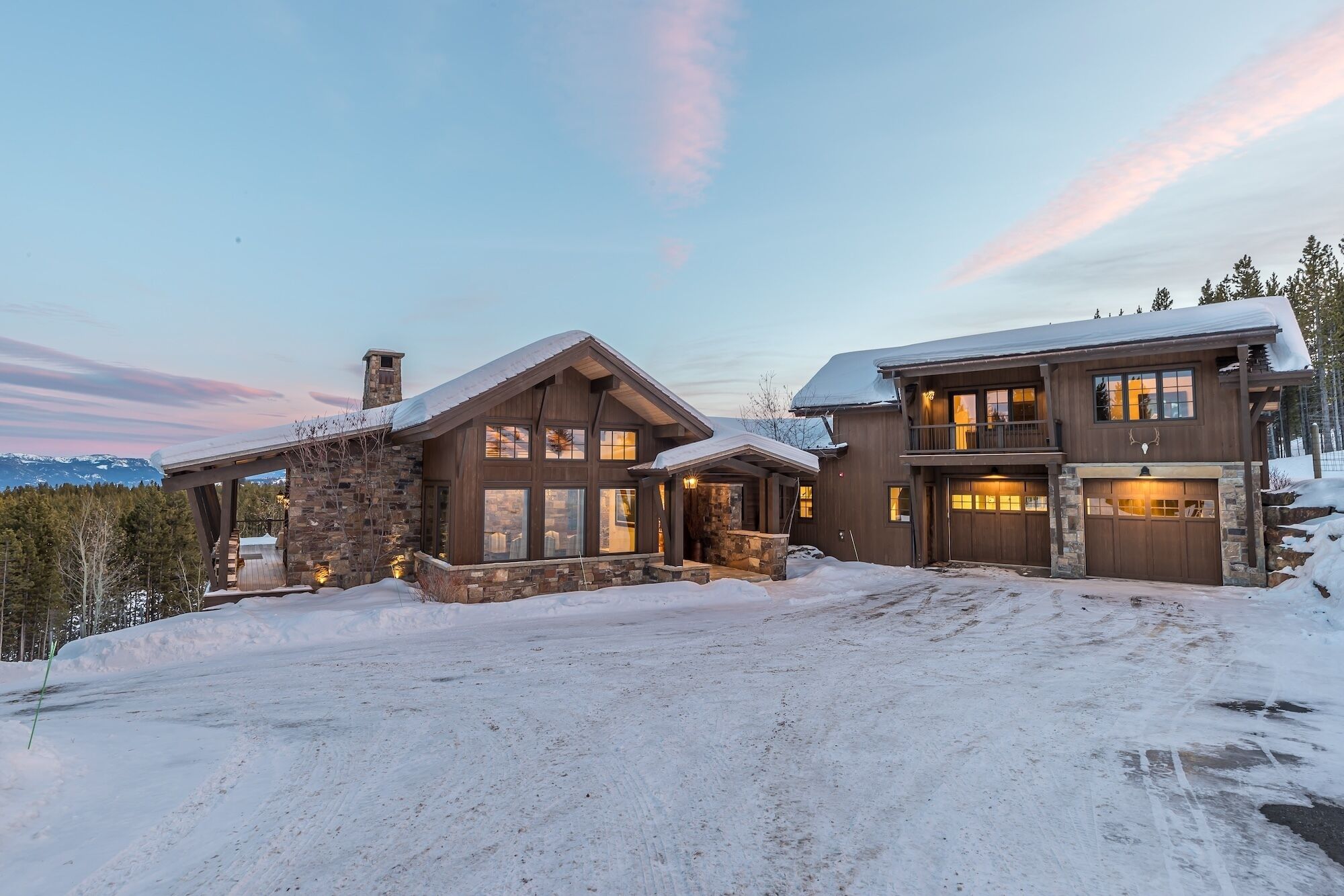 A large, two-story log cabin with a stone foundation and a snow-covered driveway.