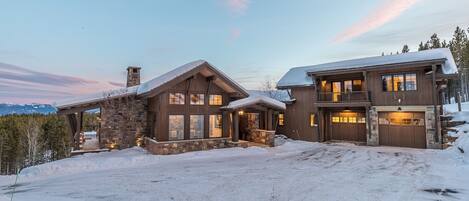 A large, two-story log cabin with a stone foundation and a snow-covered driveway.