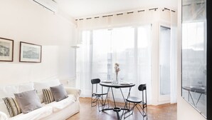 Een lichte woonkamer met een witte bank, een ronde tafel en zwarte stoelen. Er zijn grote ramen met dunne gordijnen, een uitzicht op gebouwen en ingelijste foto’s aan de muur.