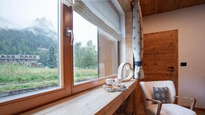 Ein gemütlicher Raum mit einer hölzernen Bank an einem Fenster mit Blick auf einen Wald und ein Gebäude. Ein Sessel mit Kissen ist ebenfalls zu sehen.