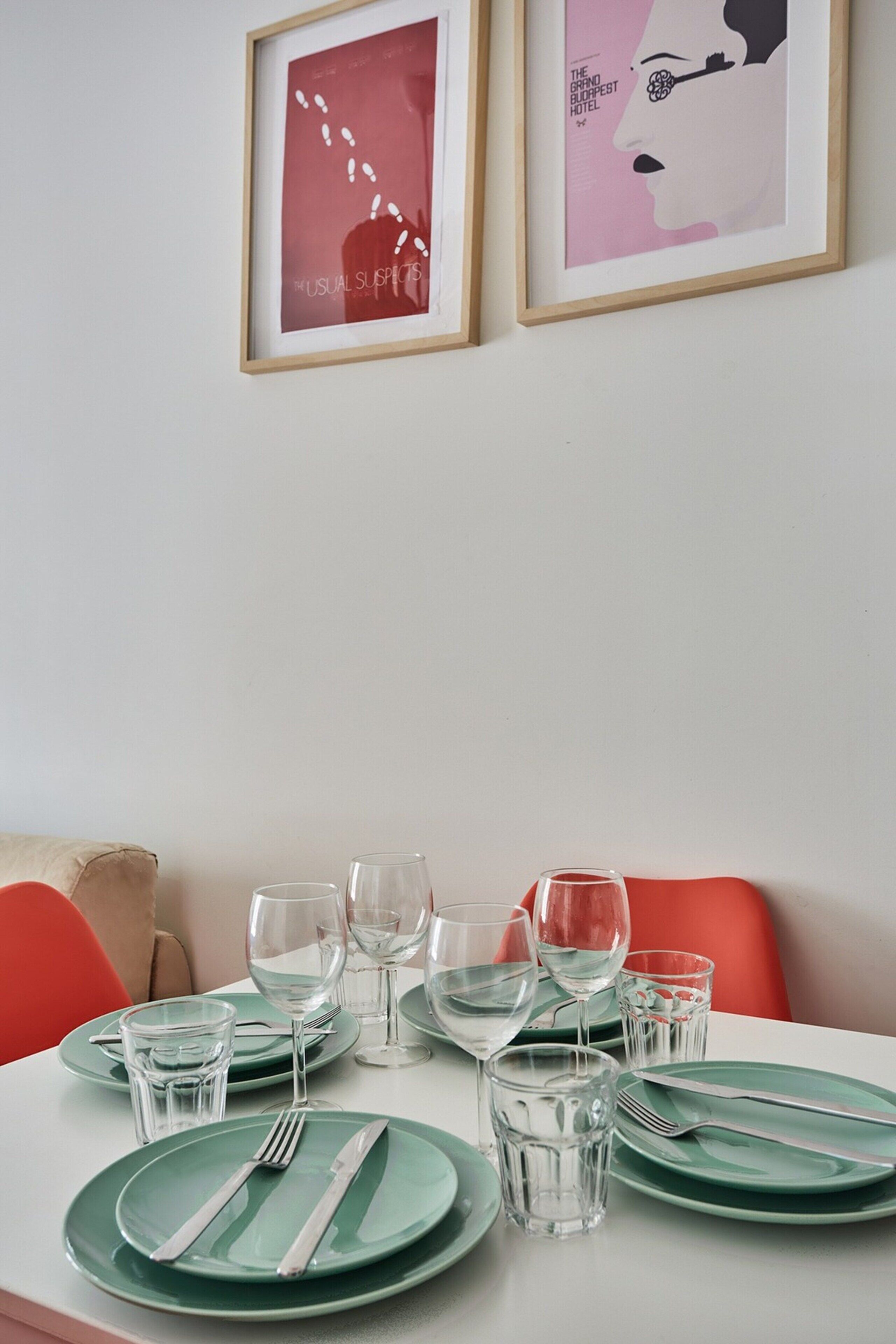 Une table à manger dressée avec de l’assiette verte, des couverts en argent et des verres transparents, une œuvre d’art encadrée au mur et une chaise rouge.