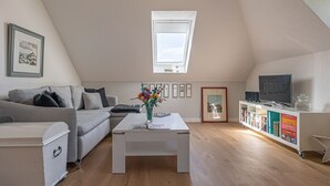 Un salón estilo loft con techo inclinado, un sofá blanco, una mesa de centro, una estantería y un televisor.