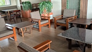 A modern indoor seating area with wooden tables and chairs, potted plants, and a view of greenery through large windows. - Hotel Sunbright Residency - Manipal (Udupi)