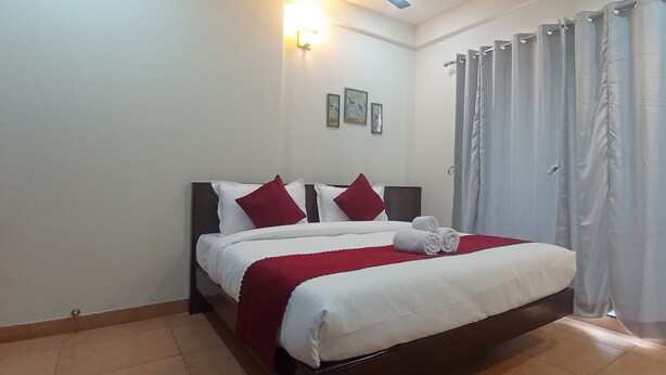 A neatly made bed with white and red bedding, a red throw blanket, and rolled towels. There are three framed pictures on the wall above the bed. - Hotel Sunbright Residency - Manipal (Udupi)