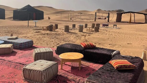 Un décor désertique avec une grande tente noire, une table en bois et plusieurs poufs disposés sur un tapis rouge.