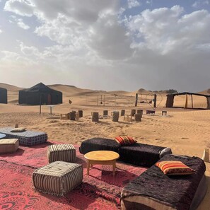 Un décor désertique avec une grande tente noire, une table en bois et plusieurs poufs disposés sur un tapis rouge.