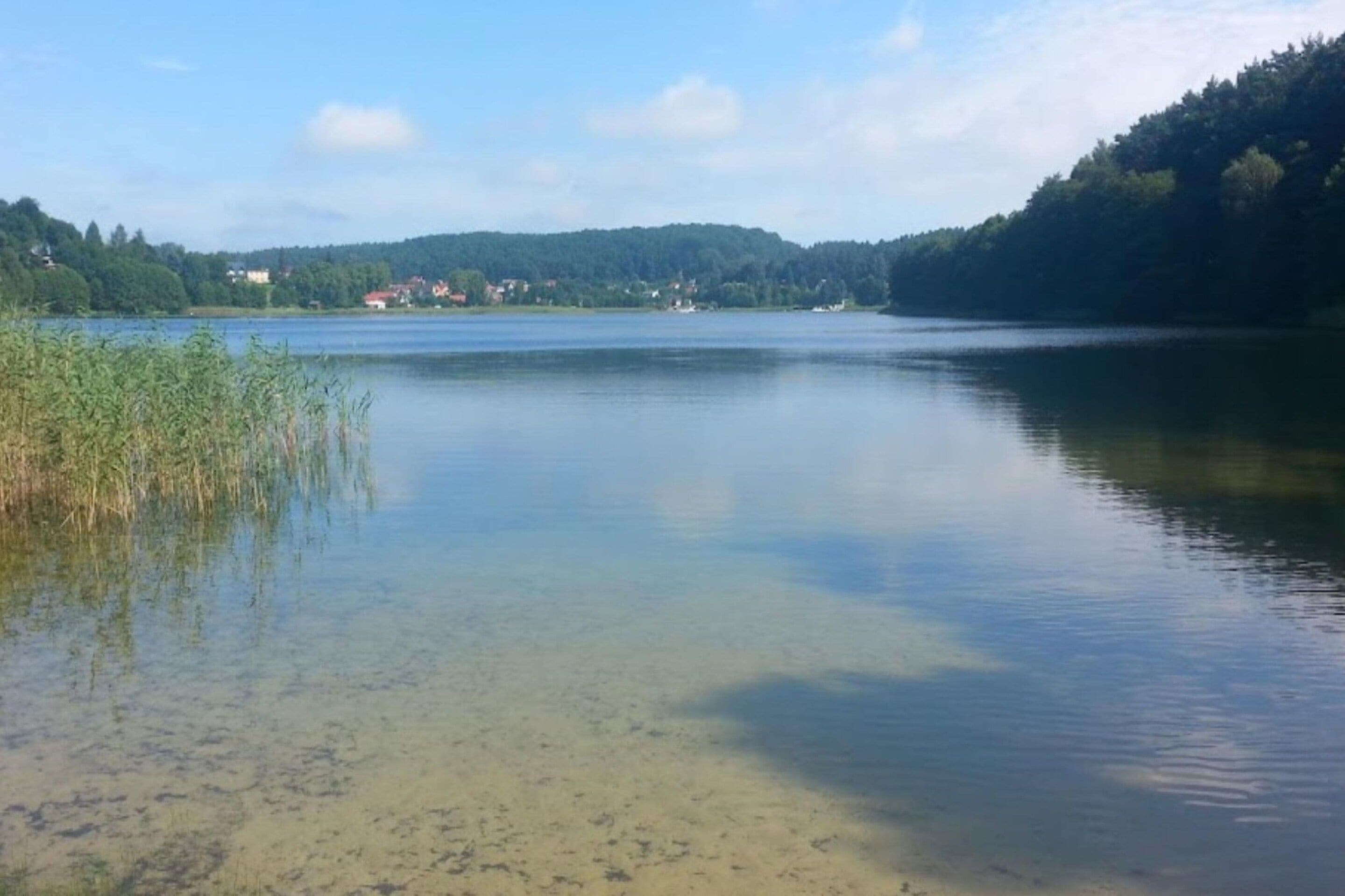 Een kalm meer met helder water, omgeven door beboste heuvels en enkele gebouwen in de verte.
