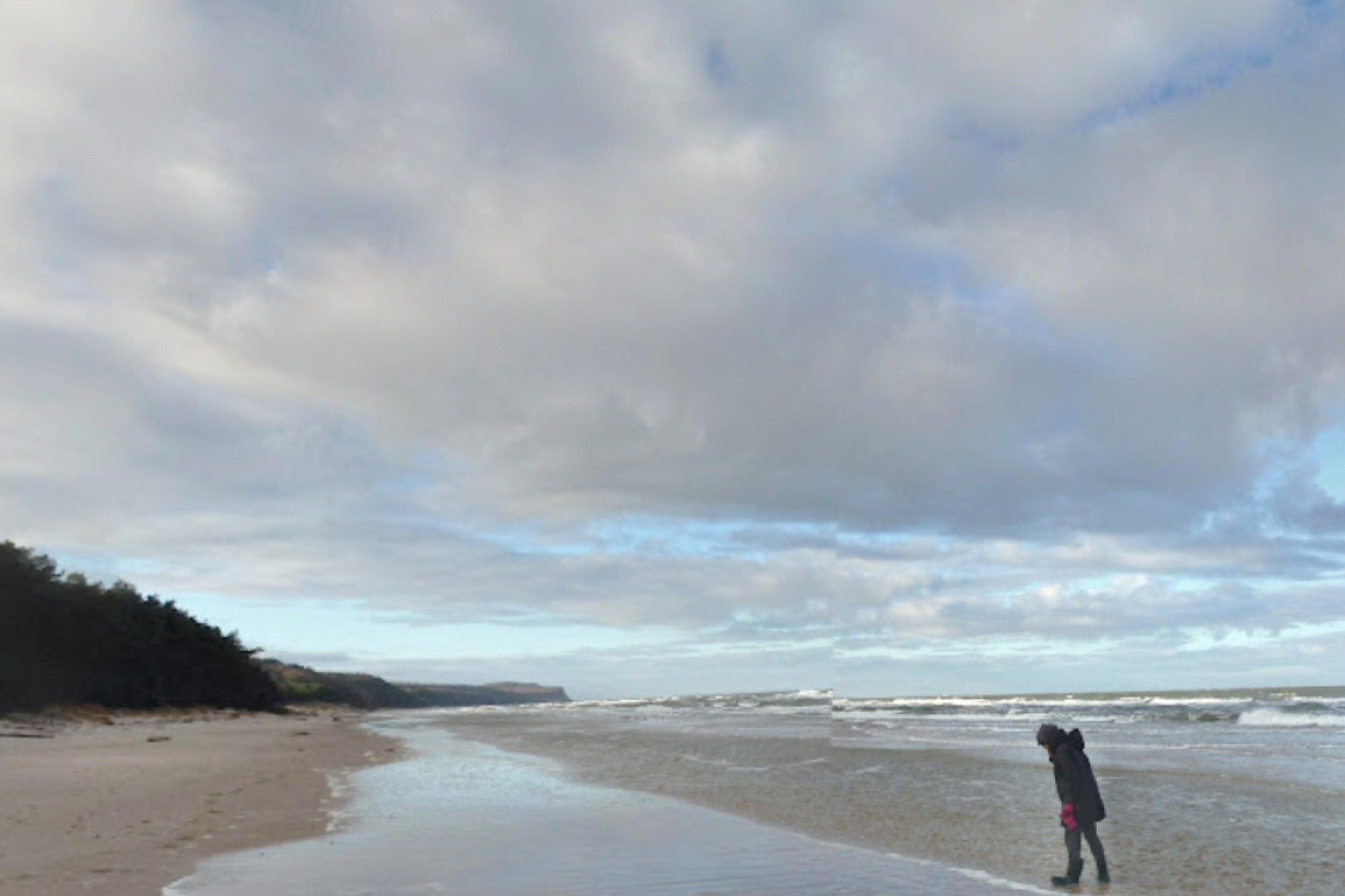 A beach with a person standing in the water, a sandy shore, and a cloudy sky.