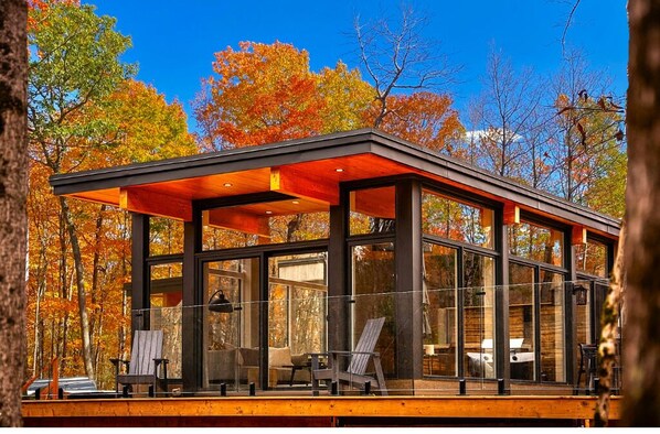 Una casa moderna de vidrio con terraza de madera, rodeada de árboles otoñales.