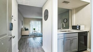 A modern kitchen with stainless steel appliances, a dining area with a sofa and a small table, and a hallway leading to a living room with a large window and a plant.