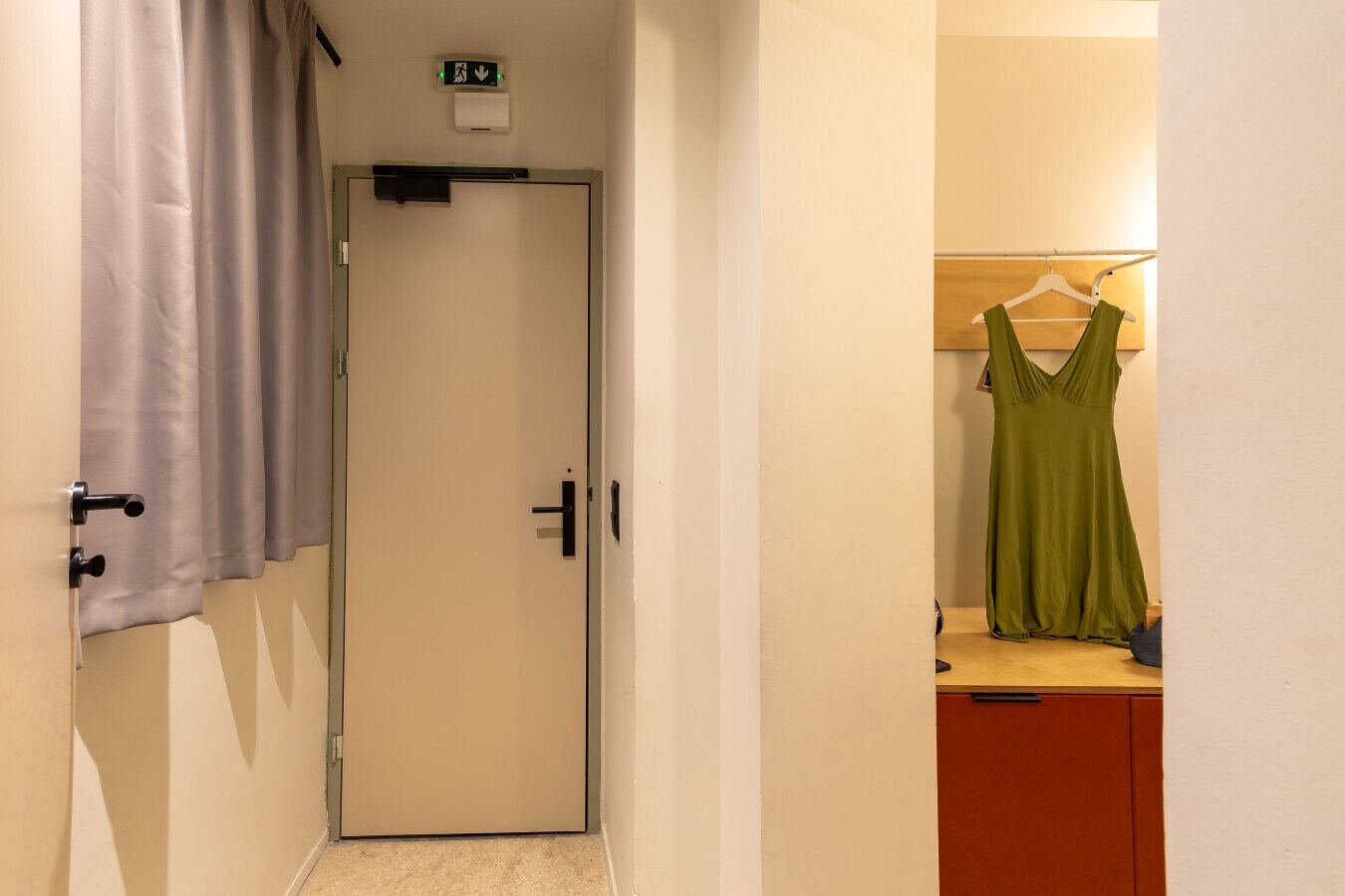 Ein Hotelzimmer mit geschlossener Tür, einem kleinen Fenster, einem Lichtschalter, einer Wand-Klimaanlage und einem grünen Kleid, das an einem Holzaufhänger hängt.