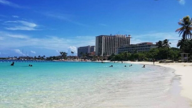 Een strand met helder turquoise water, een zandstrand en een hoog hotel op de achtergrond.