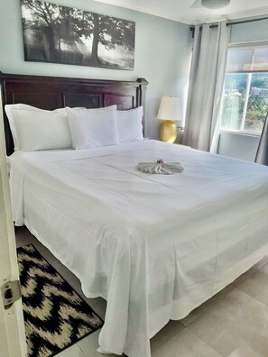 A neatly made bed with white linens, a headboard, a bedside lamp, and a view of the outdoors through a window.