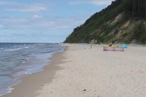 A beach with a sandy shore, a small boat, and a forested cliff in the background. - Holiday Home by Lake Wiselka, Near Baltic Beach (gmina Wolin)