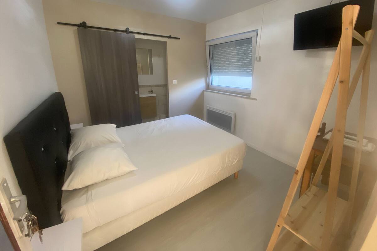 A bedroom with a bed, a wooden ladder, a TV mounted on the wall, a window with blinds, and a bathroom visible through an open door.