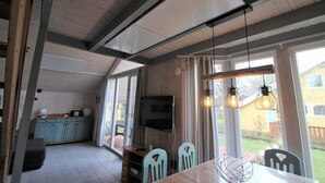 A dining area with a wooden table, chairs, and pendant lights. A television is mounted on the wall, and there is a kitchen area with cabinets and a countertop visible in the background. - Ferienpark Mirow in Granzow (Mirow)