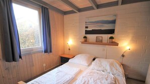 A bedroom with a wooden ceiling, a bed with white bedding, a window with curtains, a wall-mounted shelf with decorative items, and two wall-mounted lights. - Ferienpark Mirow in Granzow (Mirow)