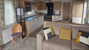 A compact kitchen with wooden cabinets, a microwave, stove, and sink. A grey sofa with yellow and black cushions is in the living area. - Shadow Escapes, Shadow Escapes - Morning Rising (Ladybank)