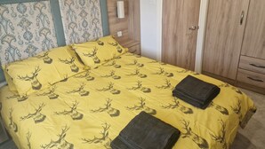 A bed with a yellow bedspread featuring a deer pattern, a wooden headboard, a bedside table with a lamp, and a wardrobe with a mirror. - Shadow Escapes, Shadow Escapes - Morning Rising (Ladybank)