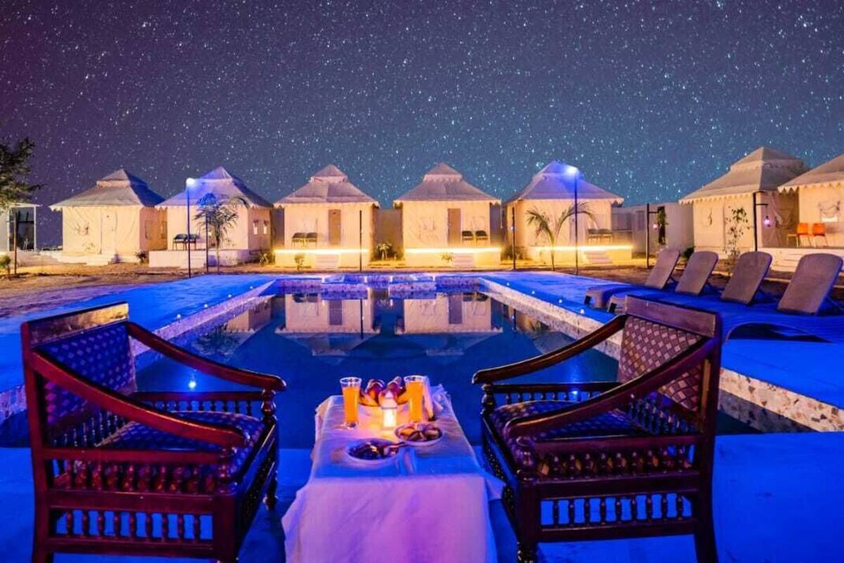 A nighttime resort with a swimming pool, lounge chairs, and tents under a starry sky.