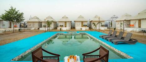 A pool area with lounge chairs and a table set for a meal, surrounded by tents.