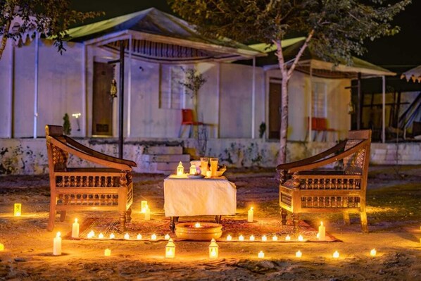 Une table et deux chaises en bois, disposées pour un repas, entourées de bougies, devant un bâtiment doté d’un porche abrité.