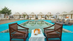 A pool area with two wooden chairs and a table set with food and drinks, surrounded by tents. - Desert Stay Camping Jaisalmer (Jaisalmer)