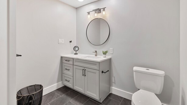 A bathroom with a white vanity, a round mirror, a toilet, and a wicker basket.