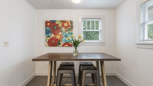 Ein Essbereich mit einem Holztisch, vier Metallhocker, einem floralen Gemälde an der Wand und einem Fenster mit Jalousien.