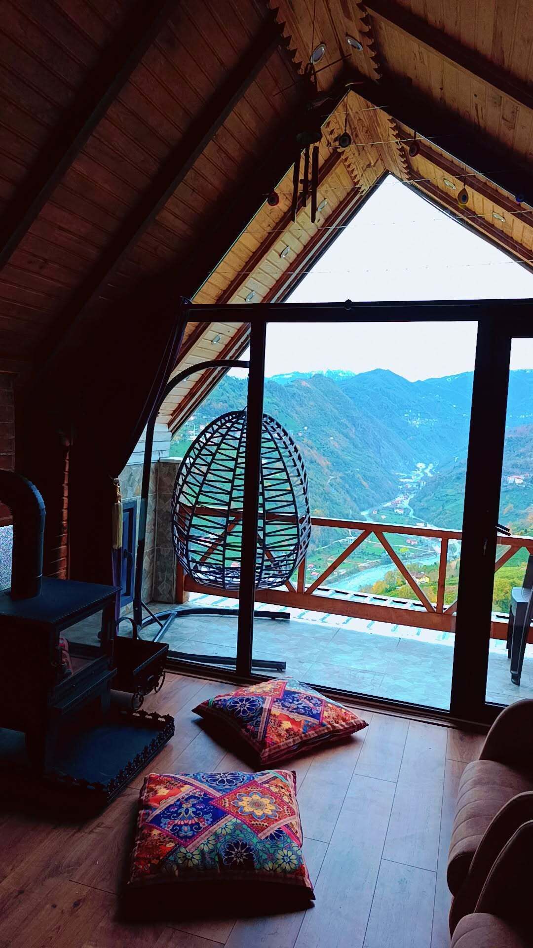 Una habitación con techo de madera, un ventanal y vistas a montañas. Hay dos cojines coloridos en el suelo y una silla colgante.