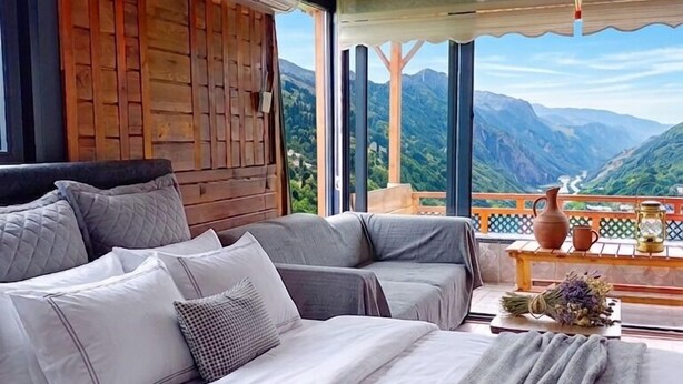 Een slaapkamer met een groot bed, een houten hoofdbord, uitzicht op bergen en een balkon met een tafel en decoratieve objecten.