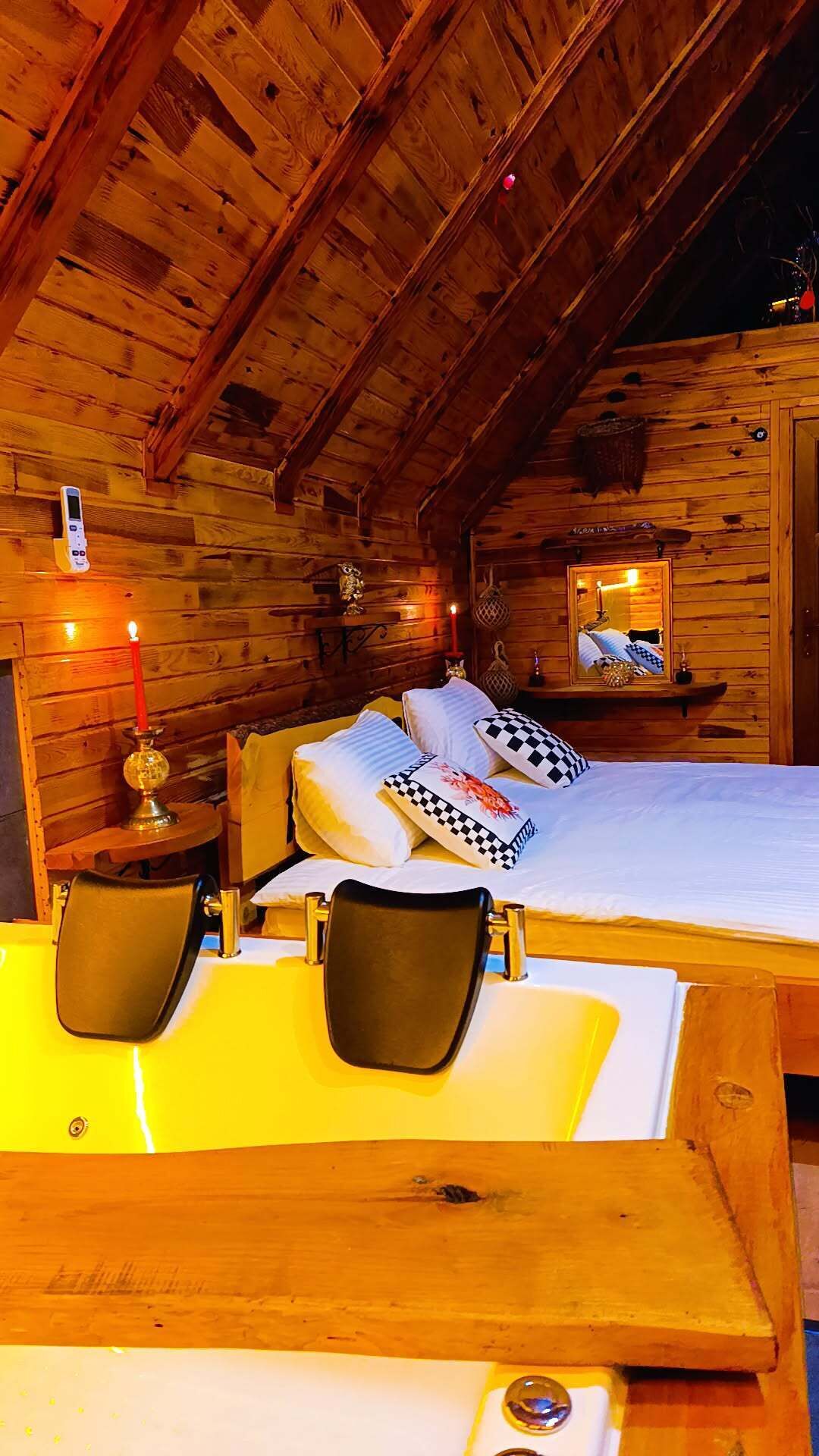 A cozy wooden cabin bedroom with a bed, two chairs, a bedside table with a candle, and a mirror on the wall.