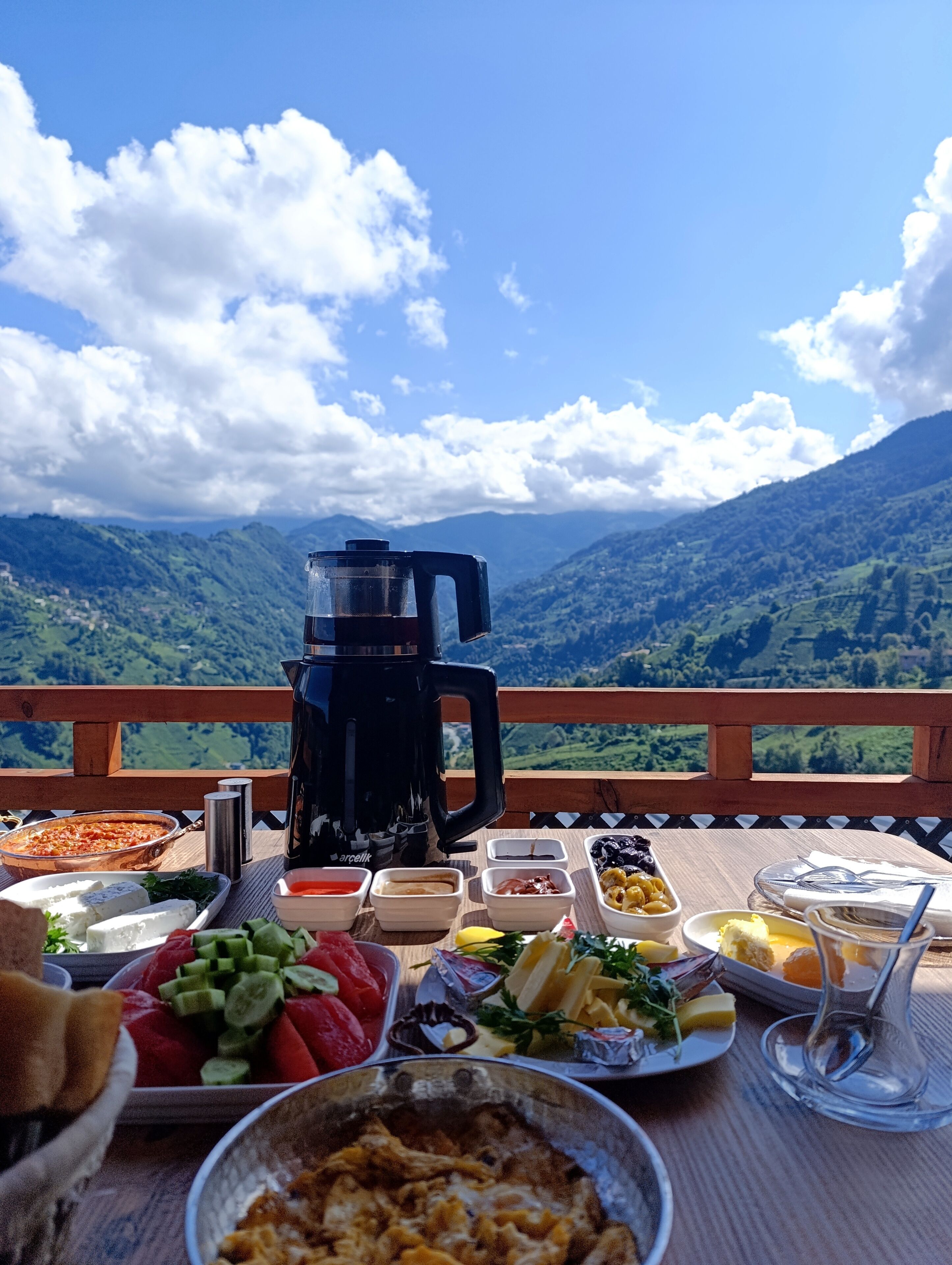 Una mesa con diversos alimentos y una cafetera, con una vista panorámica de montañas al fondo.