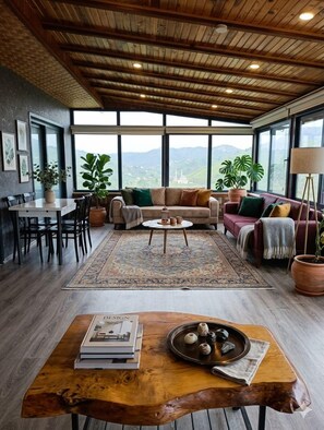 A spacious living area with a wooden ceiling, large windows, a sofa set, a dining table, and a coffee table.