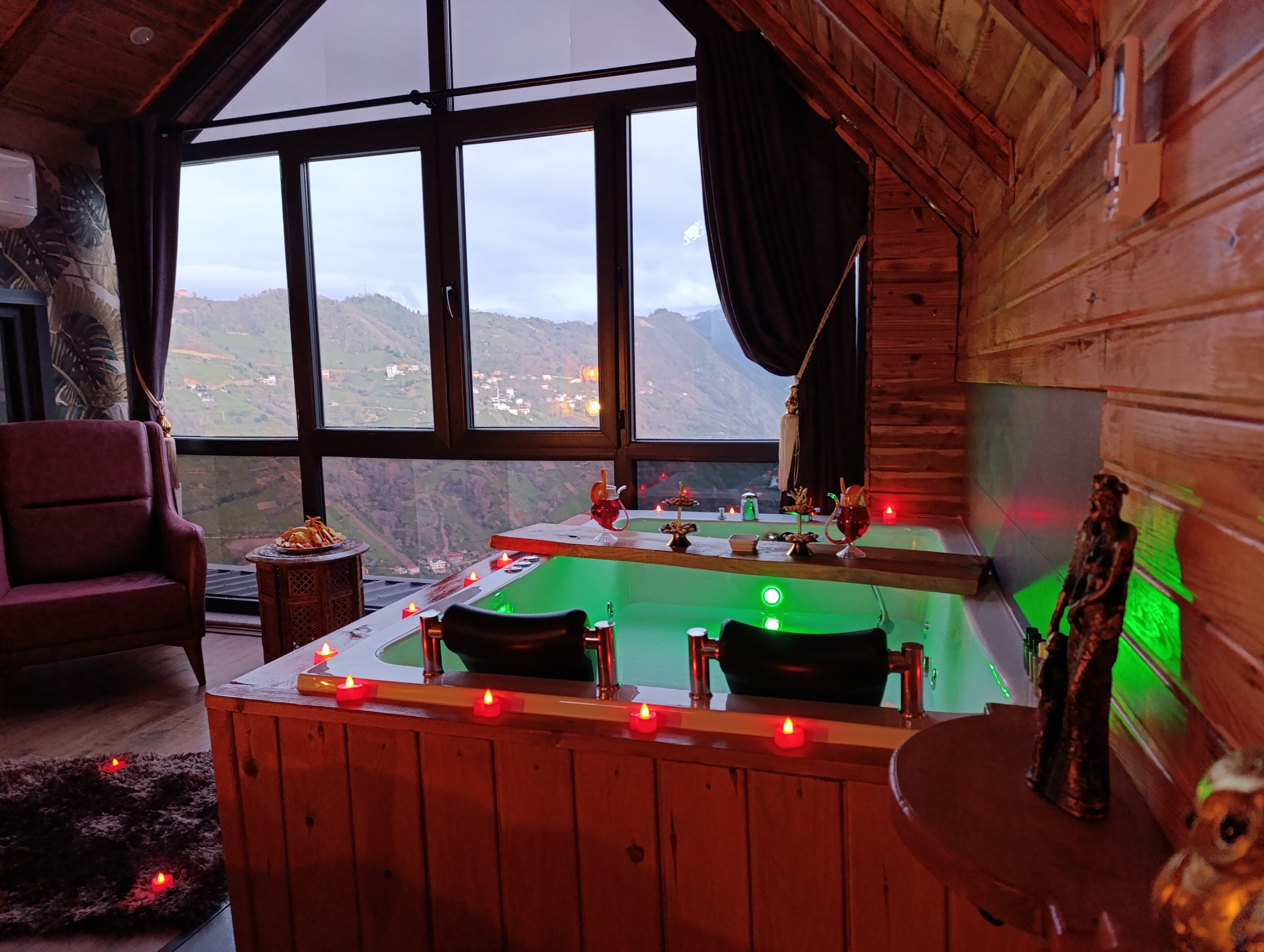 A cozy room with a wooden interior, a large window offering a mountain view, a bathtub with candles, and a plush chair.