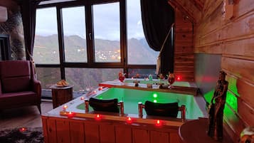 A cozy room with a wooden interior, a large window offering a mountain view, a bathtub with candles, and a plush chair.