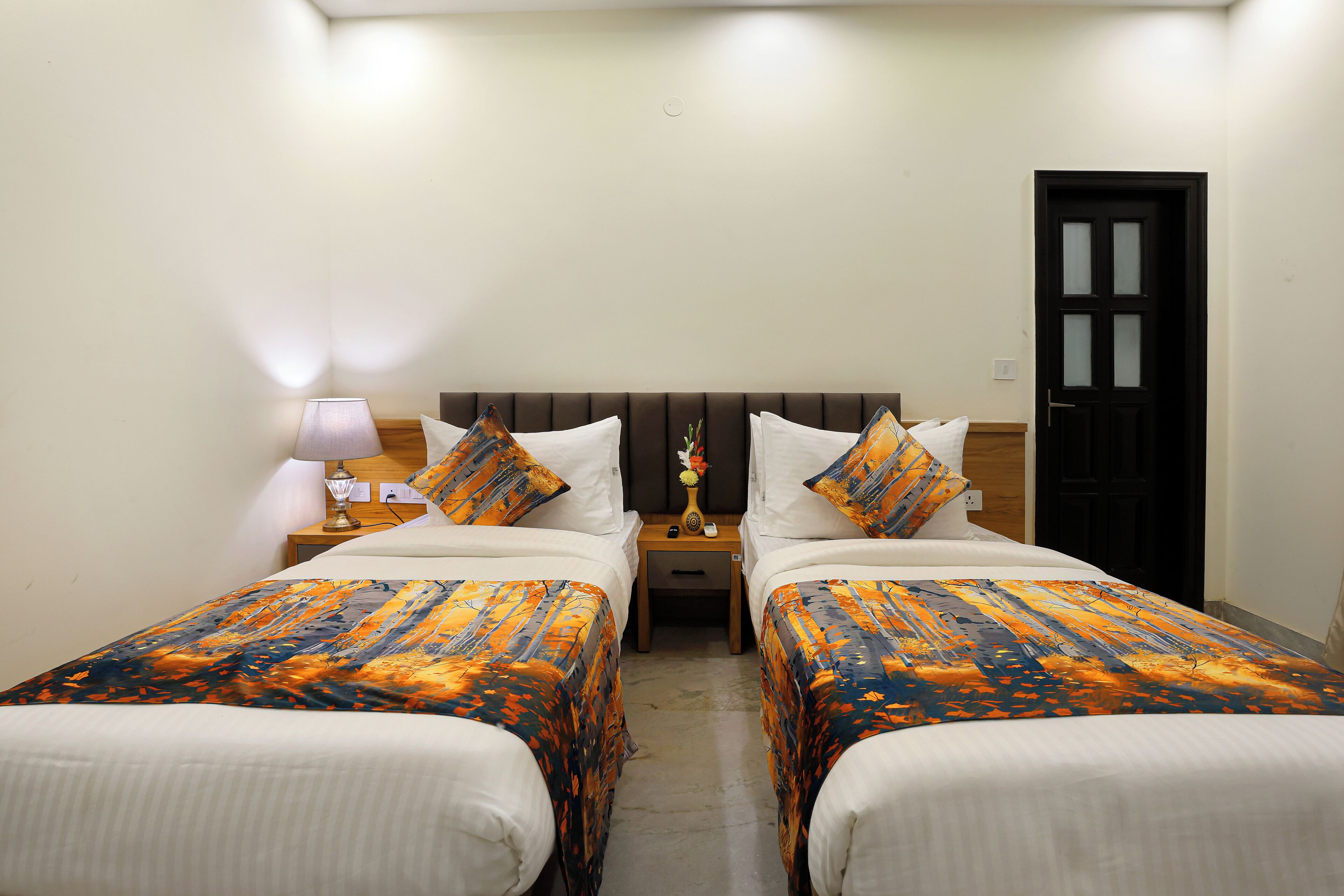 A hotel room with two beds, a nightstand with a lamp, and a door.