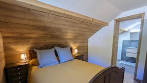 A bedroom with a wooden ceiling, a bed with pillows, a nightstand with a lamp, and a view of a bathroom through an open doorway. - Rural cottage in Vosges, 6 pers., WiFi, parking, bikes welcome (Fresse-sur-Moselle)