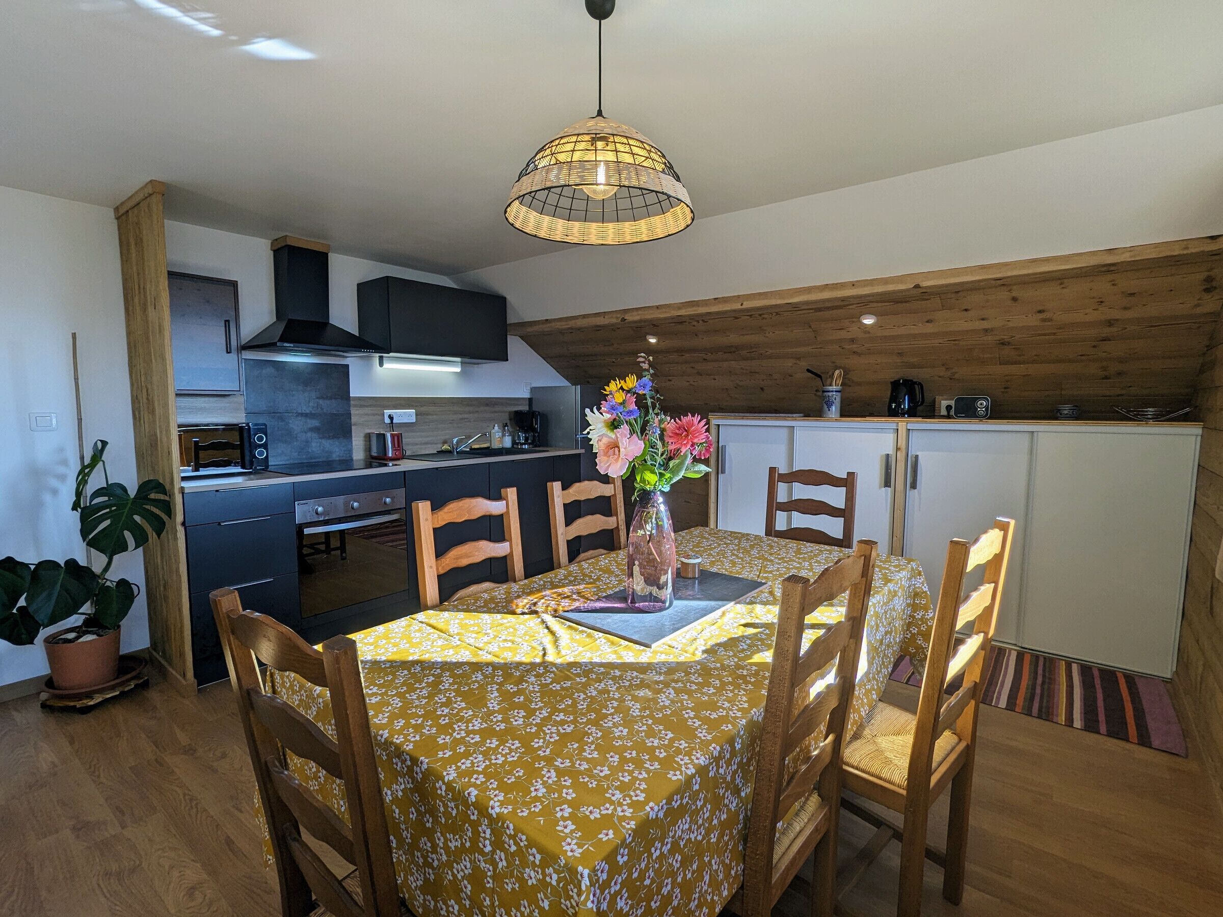 Un comedor con una mesa dispuesta para cuatro personas, rodeada de sillas de madera, un jarrón con flores y un mantel floral amarillo. Se distingue una cocina con electrodomésticos modernos y techo de madera al fondo.