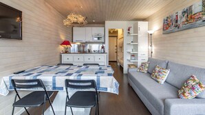 A modern kitchen with white cabinets, a dining table with chairs, a grey sofa with colorful cushions, and a ceiling with wooden slats.
