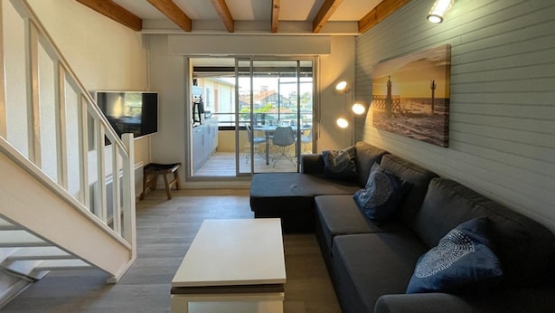 Una sala de estar moderna con un sofá, una mesa de centro y un televisor. Se observa una escalera a la izquierda y un balcón con vistas.