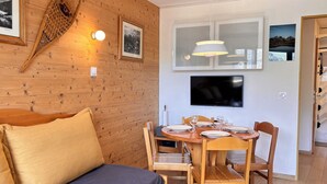 Una habitación acogedora con una pared de madera, una cama con colchón azul, una mesa de comedor preparada para dos personas con sillas, un televisor montado en la pared y cuadros enmarcados.